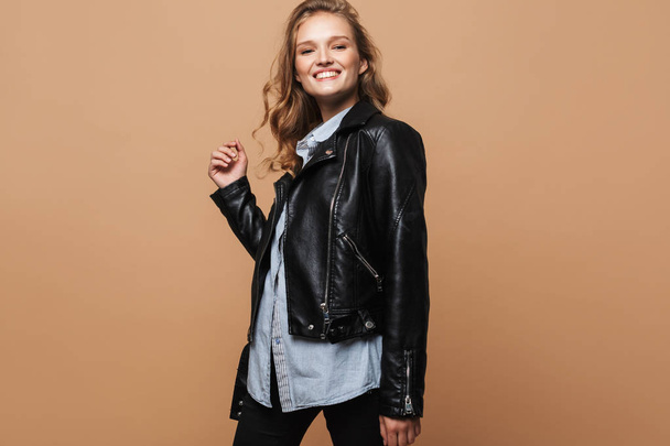 Young pretty cheerful lady with wavy hair in black leather jacket and shirt happily looking in camera over beige background - Fotografie, Obrázek