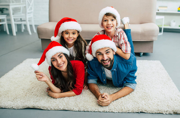 Happy young family in red santa hats spending time in living room - Zdjęcie, obraz