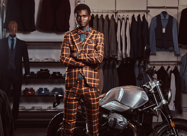 Hombre afroamericano vestido con un elegante traje de moda posando con los brazos cruzados cerca de la moto deportiva retro en la tienda de ropa para hombre
. - Foto, imagen