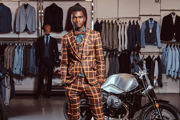 Afrikkalainen-amerikkalainen mies pukeutunut trendikäs tyylikäs puku poseeraa kädet taskussa lähellä retro urheilu moottoripyörä miesten vaatekauppa
. - Valokuva, kuva