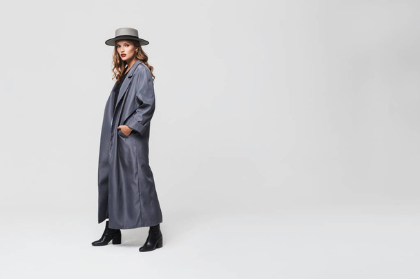 Young attractive woman with wavy hair in hat and cloak holding hands in pockets thoughtfully looking in camera over gray background - Photo, Image