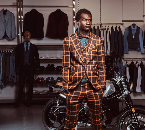 Hombre afroamericano vestido con un elegante traje de moda posando con las manos en bolsillos cerca de la moto deportiva retro en la tienda de ropa para hombre
. - Foto, imagen