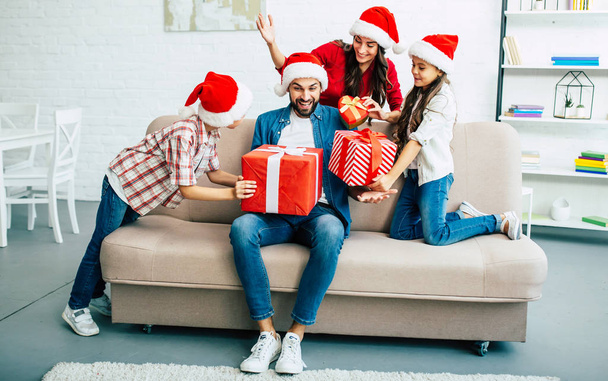 Cheerful family in santa hats giving christmas presents in living room - Photo, Image
