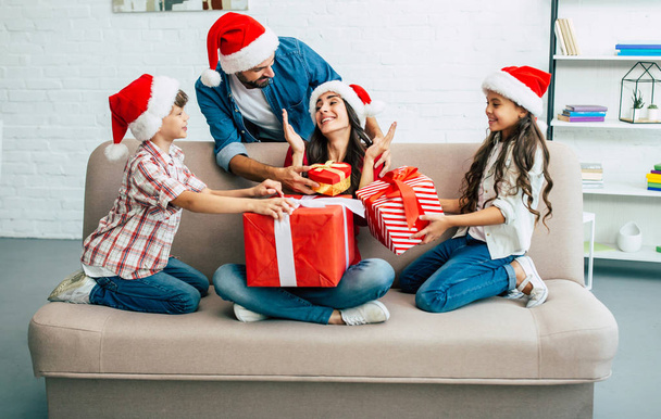 Cheerful family in santa hats giving christmas presents in living room - Fotoğraf, Görsel