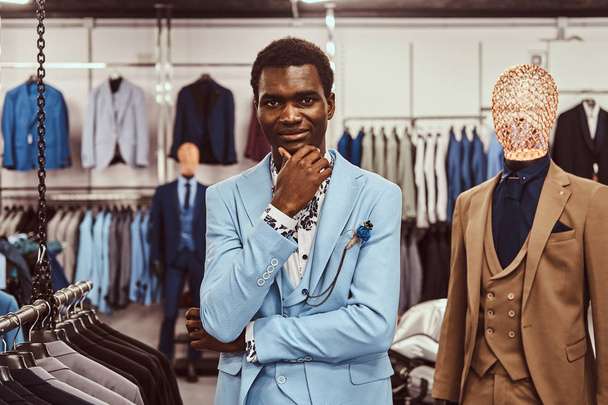 Elegancko ubrany mężczyzna Afryki pozowanie ręką na podbródek stojąc w sklepie klasycznej odzieży męskiej. - Zdjęcie, obraz