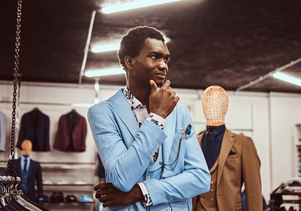 Tyylikkäästi pukeutunut Afrikkalainen mies poseeraa käsi leualla seisoessaan klassisessa miesten vaatekaupassa
. - Valokuva, kuva