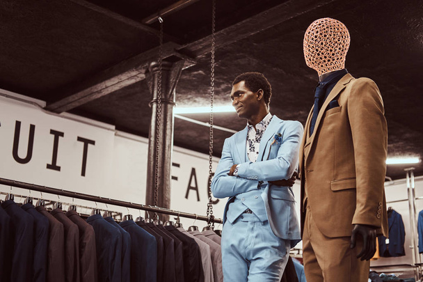 Hombre afroamericano elegante vestido posando con brazos cruzados cerca del maniquí en una tienda de ropa masculina clásica
. - Foto, Imagen