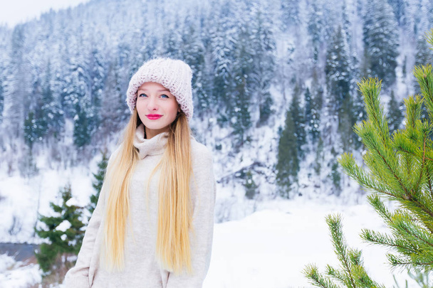 beautiful girl in a sweater and cap on her head looking at the camera lens against a snow-covered mountain - Photo, Image