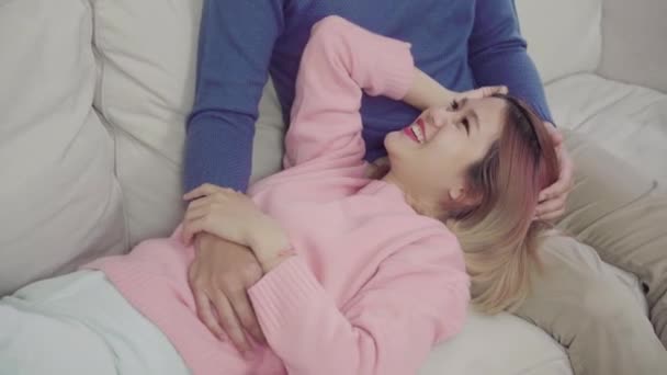 Portrait of happy sweet Asian couple have romantic time lying on the sofa when relax in their living room at home. Lifestyle couple using relax time at cozy home concept. - Video, Çekim