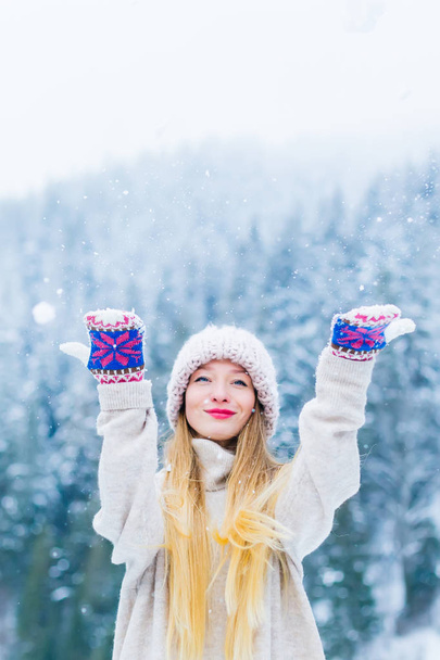 κορίτσι σε ένα πουλόβερ και καπέλο κοιτάζει το φακό της κάμερας και ρίχνει χιόνι αντιμέτωποι με φόντο τα βουνά και τα δάση - Φωτογραφία, εικόνα