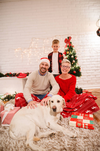 Młoda rodzina szczęśliwy obchodzi Boże Narodzenie, siedząc na podłodze w domu, z adorable pies Labrador Retriever - Zdjęcie, obraz