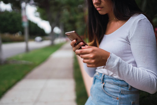 Crop young female in stylish outfit standing on sidewalk and using smartphone on blurred background of street in Los Angeles California - Фото, изображение