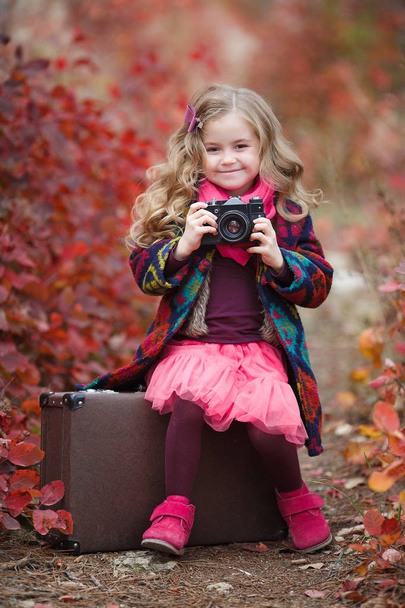 Little cute girl in an autumn jacket photographs the golden autumn, standing on the road in the woods with an old suitcase.Little kid is playing with camera and taking photo of autumn nature as master.Baby is in autumn park. - Photo, Image