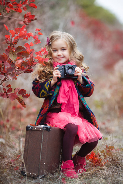 Little cute girl in an autumn jacket photographs the golden autumn, standing on the road in the woods with an old suitcase.Little kid is playing with camera and taking photo of autumn nature as master.Baby is in autumn park. - Фото, изображение