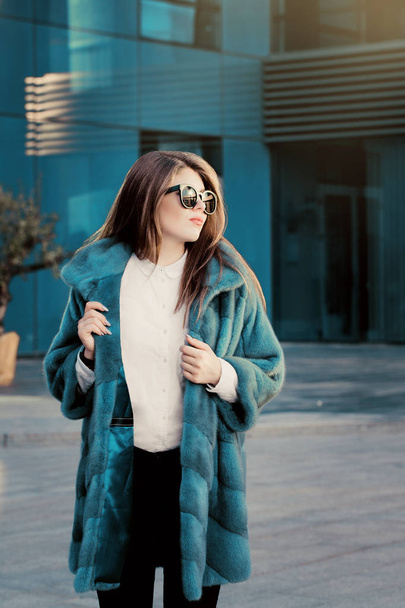 Çok genç kız beyaz gömlek, parlak renkli Doğal kürk manto ve sokakta yürürken güneş gözlüğü giyiyor - Fotoğraf, Görsel