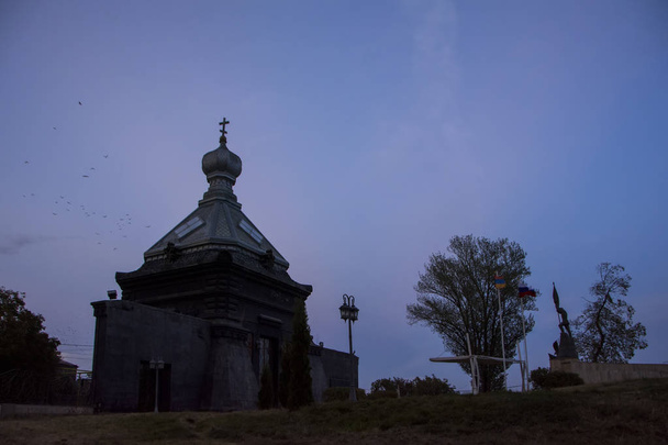 Ρωσική Ορθόδοξη Εκκλησία Αγίου Nikolai ο θαυματουργός με Αρμενικά και ρωσικά σημαίες μπροστά κάτω από τον δραματικό ουρανό ηλιοβασίλεμα στο Memorial Park, Γκιουμρί, Αρμενία. Χαμηλή γωνία προβολής - Φωτογραφία, εικόνα