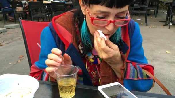 Elegante vrouw in modieuze kleding en rode bril eet vlees bij een buitencafé en gebruikt haar smartphone. Close-up. - Video