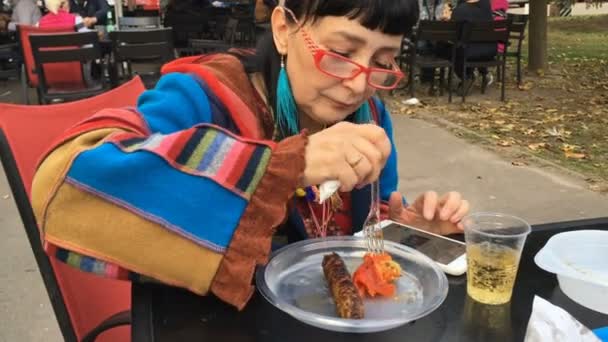 Tyylikäs nainen muodikkaissa vaatteissa ja punaisissa laseissa syö lihaa ulkona kahvilassa ja käyttää älypuhelintaan. lähikuva
. - Materiaali, video