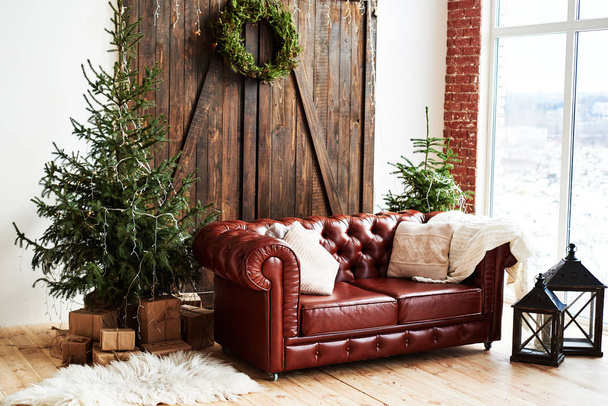 Christmas interior with vintage brown leather sofa and fir tree with gift boxes in loft room with wooden door and panoramic window, copy space - Zdjęcie, obraz