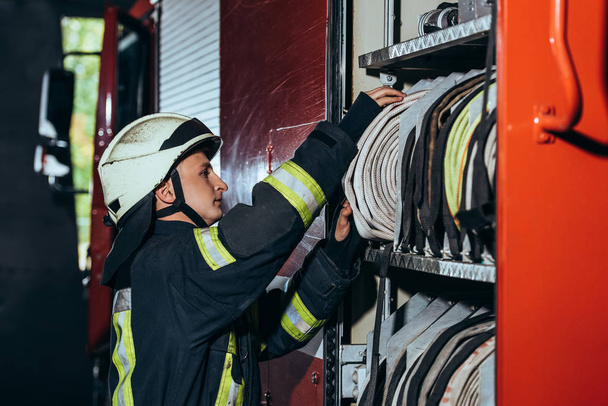 side view of fireman in protective uniform checking equipment in truck at fire station - Fotoğraf, Görsel
