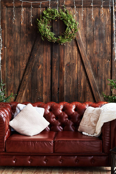 Christmas interior with vintage brown leather sofa and fir tree with gift boxes in loft room with wooden door decorated with Christmas wreath and garland, copy space - Foto, Imagem