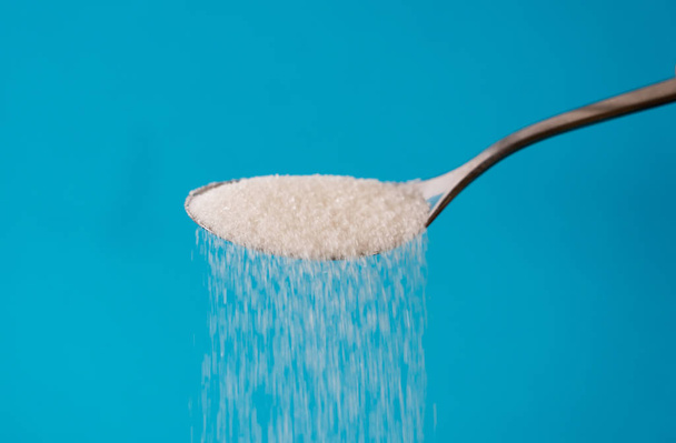 Sugar being pour from a spoon in a stream isolated on blue background in Too much sugar Sweet addiction diabetes disease unhealthy food concept. - Photo, Image