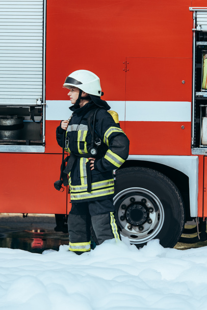 female firefighter in protective uniform standing in foam on street with red fire truck behind - Foto, afbeelding