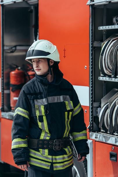 firefighter in helmet standing at truck with water hoses inside on street - Valokuva, kuva