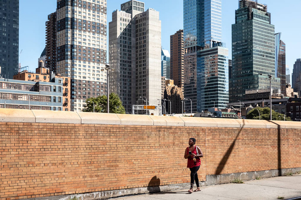 ニューヨーク、アメリカ合衆国 - Sep 23 2017: マンハッタンのストリート シーン。ニューヨーク市のマンハッタンの路上で黄色のレンガ塀に沿って携帯電話歩くと黒人の若い女性 - 写真・画像