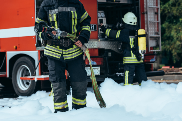 cropped shot of firefighter in water hose in hands standing in foam on street - Fotoğraf, Görsel