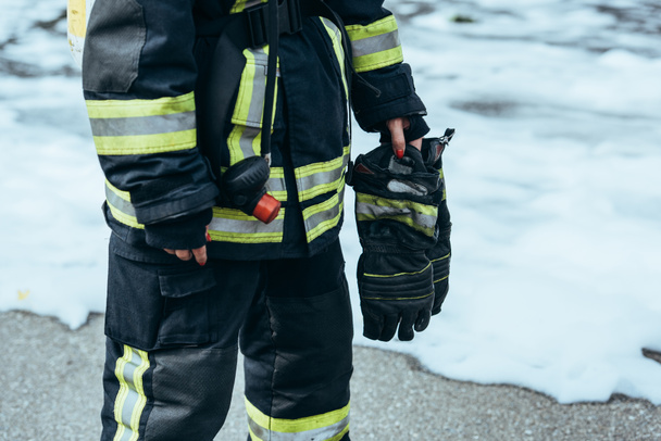 partial view of female firefighter in fireproof uniform with gloves in hand standing on steet with foam - Fotografie, Obrázek