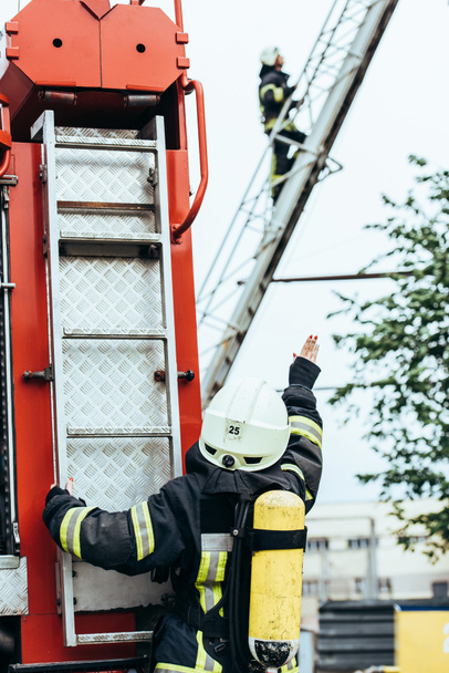 Rückansicht einer Feuerwehrfrau mit Feuerlöscher auf dem Rücken, die auf der Straße zu einem Kollegen auf der Leiter gestikuliert - Foto, Bild