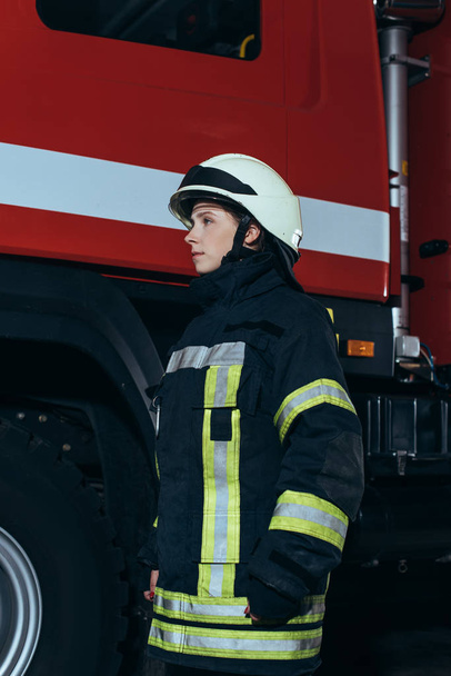 vista lateral del bombero femenino en casco de pie en la estación de bomberos
 - Foto, Imagen