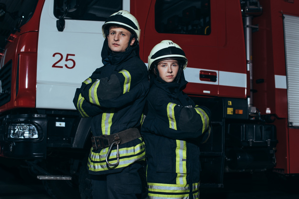 retrato de bombeiros em uniforme à prova de fogo e capacetes com armas cruzadas de pé perto do caminhão no quartel de bombeiros
 - Foto, Imagem