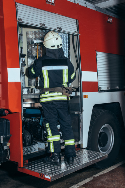 back view of firefighter in fireproof uniform and helmet standing near truck at fire station - Fotoğraf, Görsel