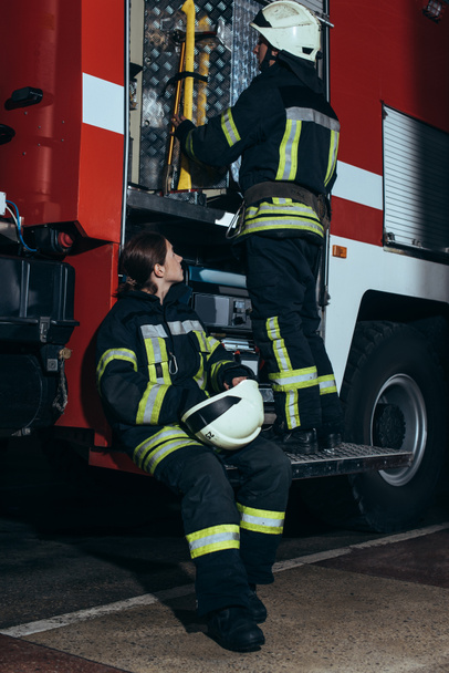 female firefighter in protective uniform looking at colleague checking equipment in truck at fire department - Foto, afbeelding