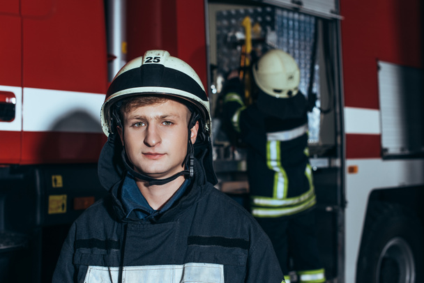 vigile del fuoco maschio in casco guardando la telecamera con collega che controlla le attrezzature dietro alla stazione dei pompieri
 - Foto, immagini