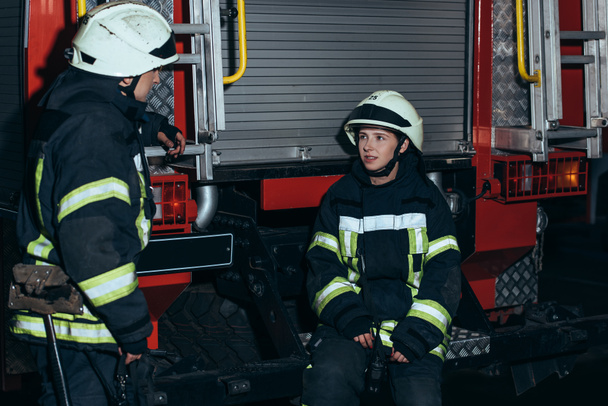 Feuerwehrleute in feuerfester Uniform und Helmen unterhalten sich am Feuerwehrhaus - Foto, Bild