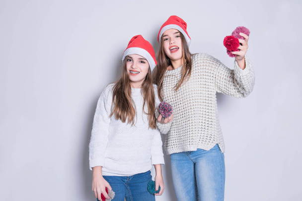 Portrait of two happy teenage girls in santa claus hat and white warm sweaters. Merry Christmas and Happy Holidays concept. Family holiday, party over white background. Cute teen sisters. - Photo, Image