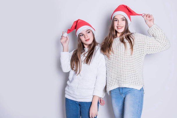 Portrait of two happy teenage girls in santa claus hat and white warm sweaters. Merry Christmas and Happy Holidays concept. Family holiday, party over white background. Cute teen sisters. - 写真・画像