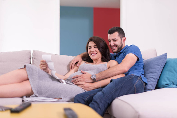 Young pregnant couple looking baby's ultrasound photo while relaxing on sofa at home - Fotoğraf, Görsel