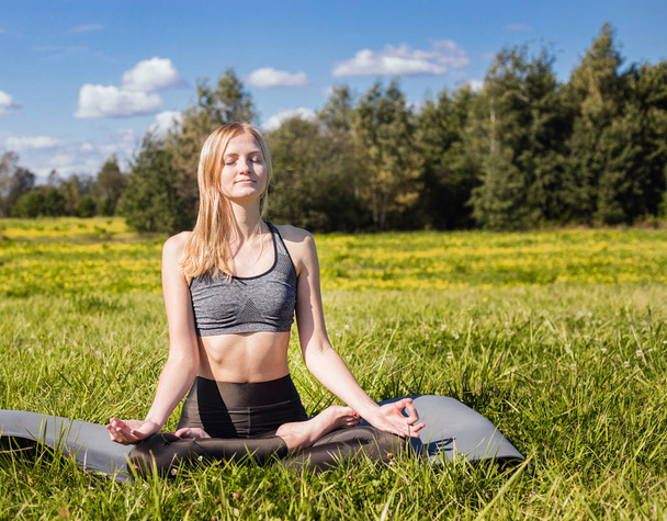 Young female with open arms and long blond hair relaxes in yoga pose in green nature - Photo, image