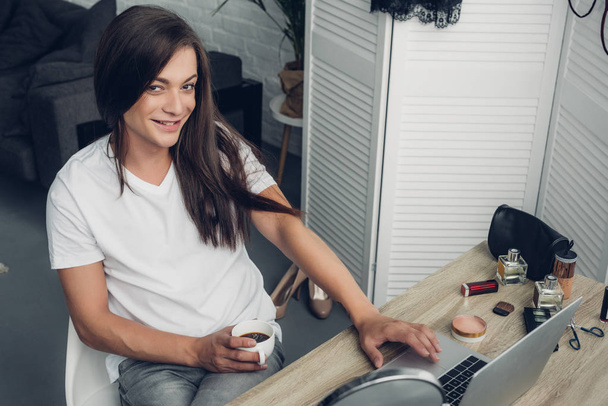 若いトランスジェンダーのフリーランスの女性を笑顔にしながら家でコーヒーを飲みながらカメラを見て - 写真・画像