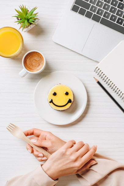 vista parcial de la mujer de negocios comiendo pastel con símbolo de sonrisa en la mesa con jugo de naranja y taza de café en la oficina
  - Foto, Imagen