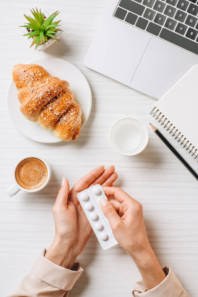 обрезанный образ деловой женщины, принимающей таблетки перед завтраком за столом с круассаном и чашкой кофе в офисе
  - Фото, изображение