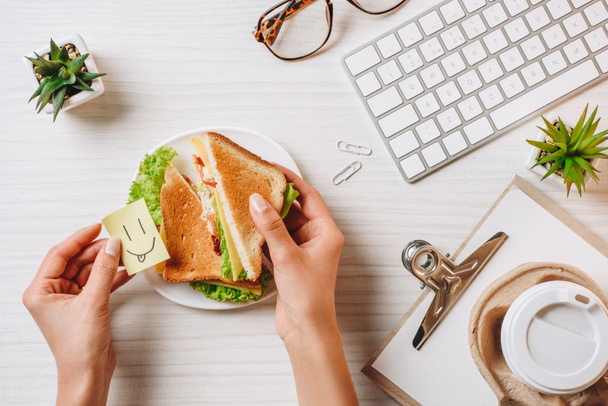 частичный вид деловой женщины с символом улыбки, обедающей с бутербродом и кофе в бумажной чашке за столом в офисе
  - Фото, изображение