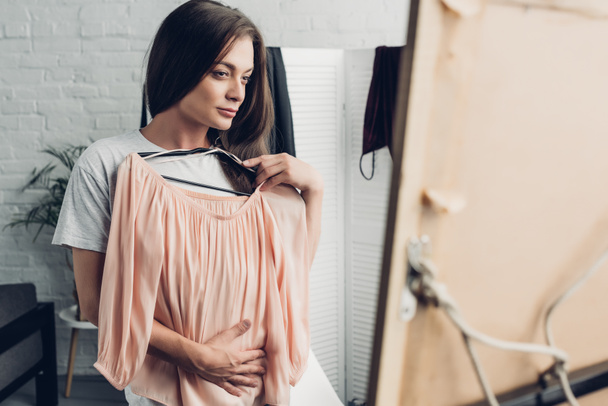 atractiva mujer transgénero probándose la camisa femenina y mirando el espejo en casa - Foto, Imagen