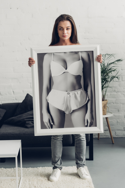 serious young transgender woman holding black and white photo with female body in underwear at home - Zdjęcie, obraz