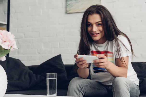 mosolygós fiatal transznemű nő használ okostelefon kanapén otthon - Fotó, kép