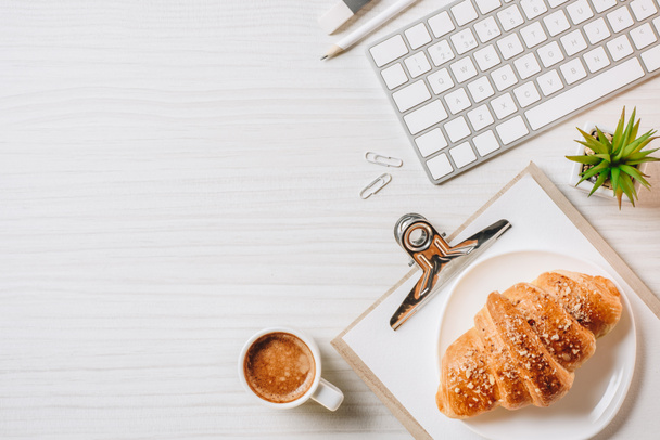 elevated view of clipboard, computer keyboard, croissant and coffee cup at table in office  - Fotografie, Obrázek